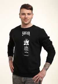 Muž s krátkými vlasy, oblečený v černé mikině s grafikou dívky a textem "Sarkasmus je jediný přítel, který potřebuji ve středu," stojící s rukama v bok.