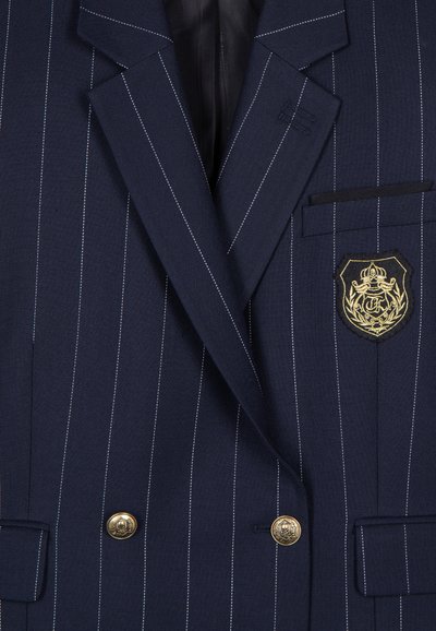 Blazer doppiopetto blu navy con righe bianche, bottoni dorati e un emblema ricamato sulla tasca del petto. Design strutturato e texture del tessuto.