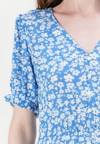 Robe bleue à motifs floraux blancs, à manches courtes bouffantes, décolleté en V et boutons sur le devant. Tissu doux et texturé.