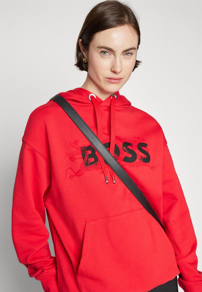 Sweat à capuche rouge avec un grand logo noir "BOSS", comportant des œillets en métal, des cordons de serrage et une poche avant. Sangle noire sur l'épaule.