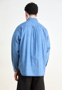 Lichtblauwe spijkerhemel met een relaxte pasvorm, lange mouwen en knoopmanchetten. Beschikt over een rugplooien voor extra comfort en bewegingsvrijheid.