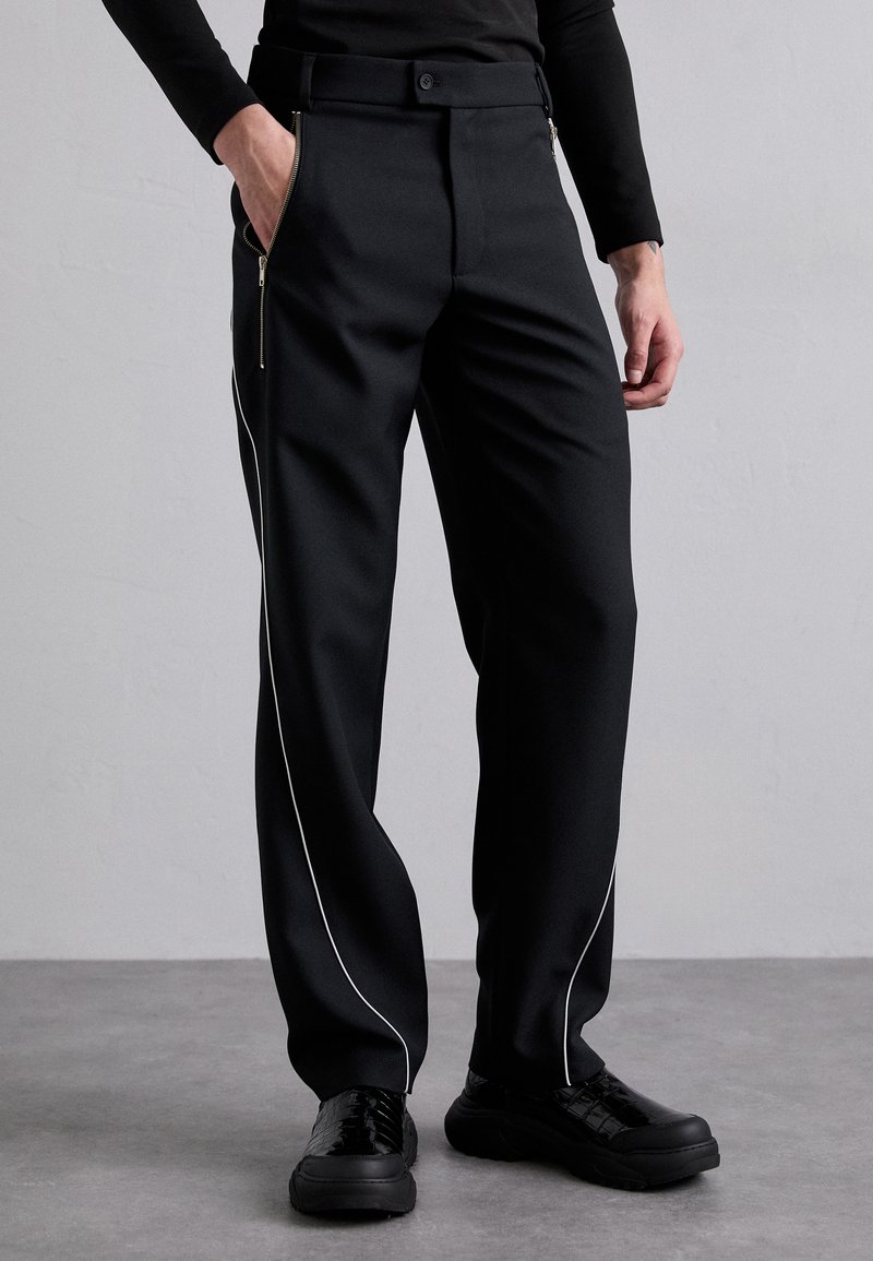 Pantalons noirs à coupe ajustée, dotés de passepoils blancs sur les côtés et de poches zippées. Fabriqués en tissu tissé, associés à des baskets noires.