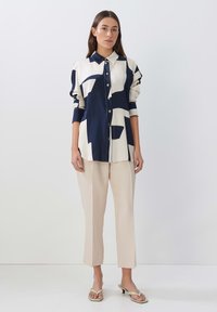 Navy and cream patterned button-up shirt with long sleeves, paired with beige tapered pants and light sandals, standing against a plain background.