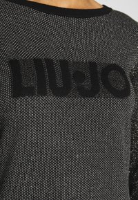 Gros plan sur un pull en tricot noir et argenté avec un motif texturé et un grand logo noir "LIU JO" sur la poitrine.