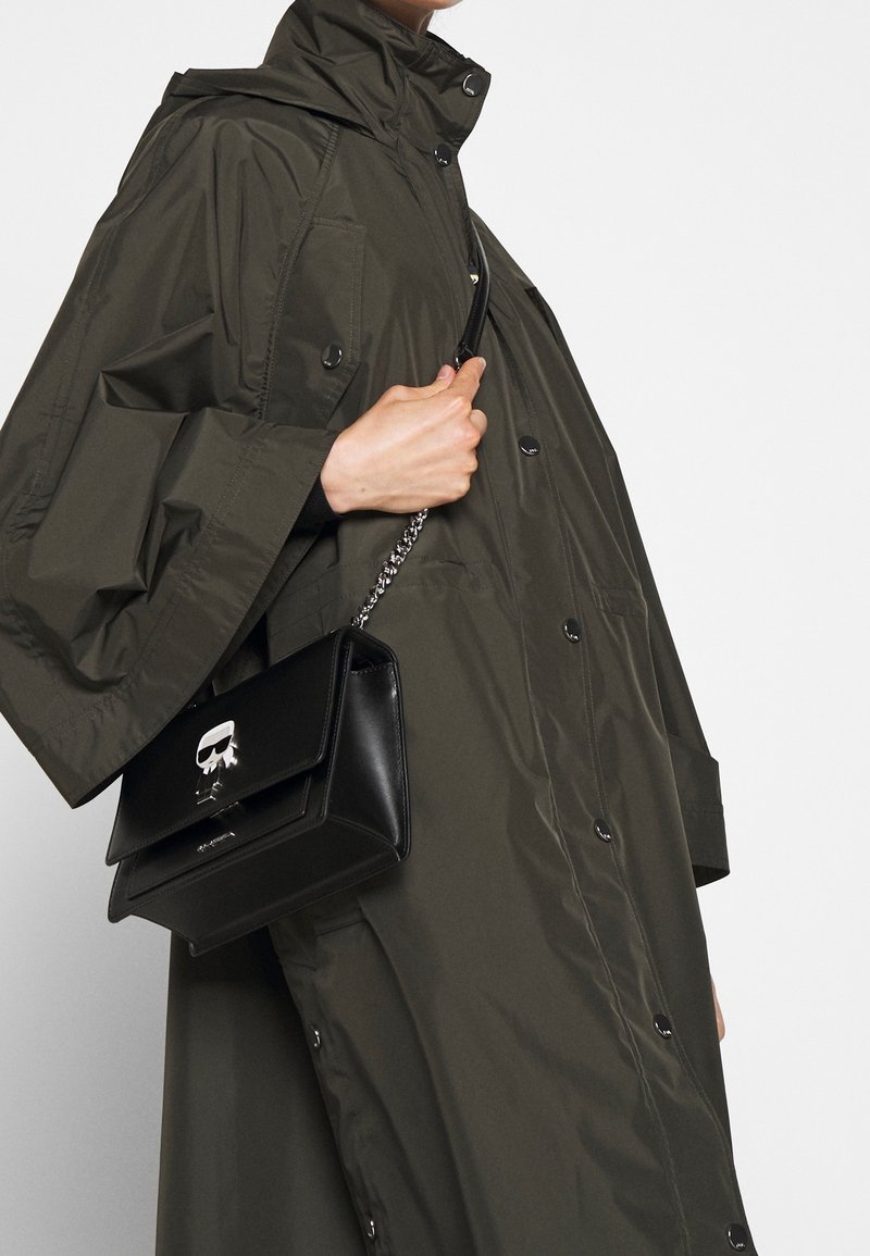 Olivi vihreä vedenpitävä takki, jossa on nepparit, yhdistettynä mustaan nahkakäsilaukkuun, jossa on hopeinen ketjuremmi ja pääkalloaksentti.