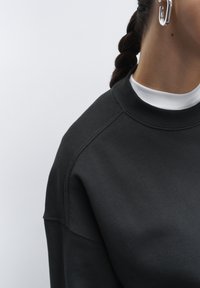 Sudadera negra con cuello redondo, puños acanalados y una textura suave. Detalles de costura visibles y se lleva capa sobre un cuello de tortuga blanco debajo.