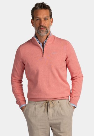 Man met grijze baard die een koraalkleurige trui met ritssluiting draagt over een blauw overhemd met kraag, beige broek, handen in de zakken, tegen een effen achtergrond.