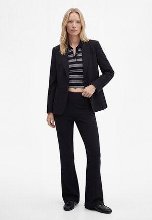 Kvinde iført sort blazer og bukser med sort-hvid stribet kort skjorte, stående mod en ensfarvet hvid baggrund.