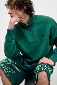 Zelená mikina so zipsom a vysokým golierom v kombinácii so zelenými šortkami s žltými grafickými akcentmi. Hladká textúra látky, voľný štýl.