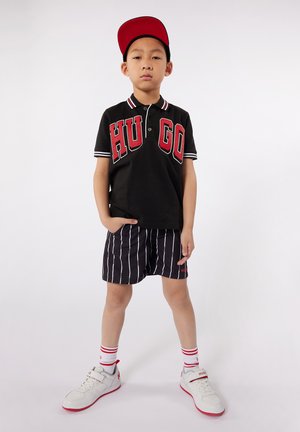 Maglietta polo nera con scritta rossa "HUGO", pantaloni corti neri a righe, scarpe da ginnastica bianche con dettagli rossi e un cappellino rosso. Design semplice, vestibilità casual.