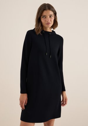 Vrouw met schouderlang bruin haar in een zwart jurk met lange mouwen en een koordsluiting bij de hals, staand tegen een beige achtergrond.