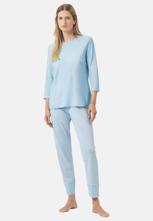 Vrouw staat blootsvoets met een lichtblauw shirt met lange mouwen en gestreepte lichtblauwe pyjamabroek tegen een witte achtergrond.