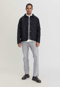 Schwarze gesteppte Jacke mit Druckknopfverschluss und Kapuze. Kombiniert mit einem grauen Pullover und hellgrauen Jeans, abgerundet mit schwarzen Schuhen.