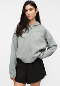 Γκρι φούτερ με φερμουάρ και άνετη εφαρμογή, διαθέτει κουκούλα, μακριά μανίκια και λογότυπο στο στήθος. Φοριέται με μαύρες βερμούδες.