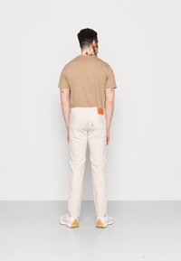 Camiseta beige de manga corta, combinada con pantalones blancos que cuentan con una etiqueta de cuero y bolsillos traseros estándar. El calzado son zapatillas deportivas blancas.