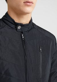 Czarna pikowana kurtka z wysokim kołnierzem, zamek błyskawiczny z przodu oraz jedną kieszenią zapinaną na zamek na klatce piersiowej. Wykonana z gładkiego, lekkiego materiału.