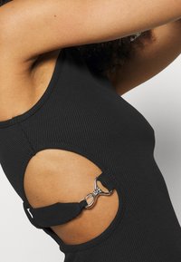 Černý žebrovaný tílko s výřezem po bocích, zdobené stříbrným kováním a detaily na ramínkách. Hladká textura a přiléhavý střih.