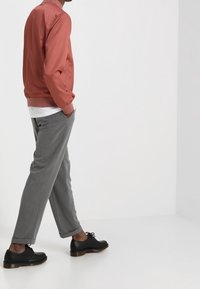 Man walking with hands in pockets, wearing a rust-colored jacket, white shirt, gray pants, and black lace-up shoes on a white background.