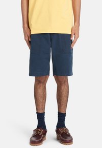 Pantaloni corti in cotone blu navy con vestibilità rilassata, dotati di due tasche frontali e taglio dritto. Indossati con scarpe da barca marroni e calze blu navy.
