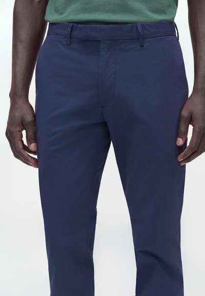 Homme portant un pantalon chino bleu foncé et une chemise verte, visible de la taille jusqu'au milieu des cuisses, debout avec les bras détendus le long du corps.