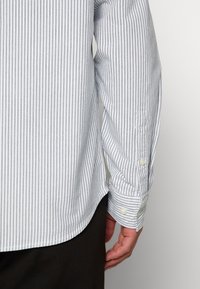 Chemise à rayures boutonnée sur le devant, présentant des rayures verticales bleu clair et blanches, avec un ourlet arrondi et des poignets boutonnés. Texture de tissu lisse.