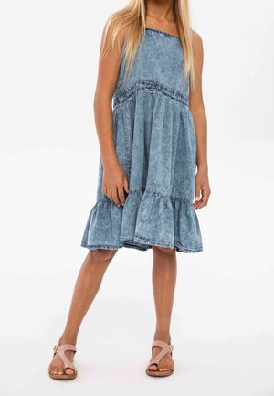 Robe en denim avec une jupe à volants, de couleur bleu clair, smocks élastiques à la taille et fines bretelles. Associée à des sandales beiges.