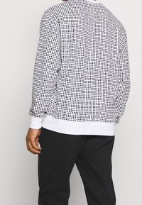Camisa de manga larga a rayas en blanco y negro con un diseño de cuadrícula. Presenta puños acanalados y un dobladillo blanco, fabricada con una tela suave.