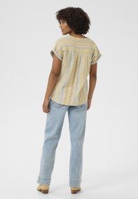 Kortärmad, mönstrad blus i gult och ljusblått; lättviktig tyg med plisserad bakdetalj. Matchad med ljus tvätt, raka jeans.