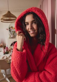 Red fluffy hooded robe with textured fabric, featuring a deep hood and wide sleeves. Background includes a decorated dining area.