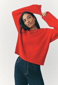 Maglione rosso a maglia con una vestibilità ampia e orlo a coste, abbinato a jeans in denim scuro. Le maniche sono lunghe e leggermente oversize.