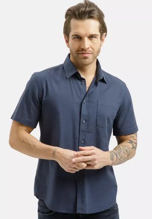 Mężczyzna z ułożonymi brązowymi włosami, ubrany w granatową koszulę z krótkim rękawem i guzikami, pokazujący tatuaże na obu przedramionach, z rękami złożonymi przed sobą.