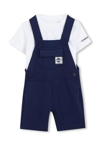 Salopette corta blu navy con tasca frontale e toppa con logo, abbinata a una maglietta bianca. Presenta bretelle regolabili e chiusura con bottoni.