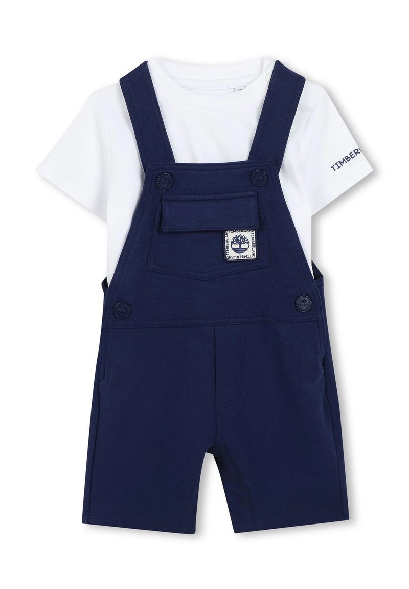 Salopette corta blu navy con tasca frontale e toppa con logo, abbinata a una maglietta bianca. Presenta bretelle regolabili e chiusura con bottoni.