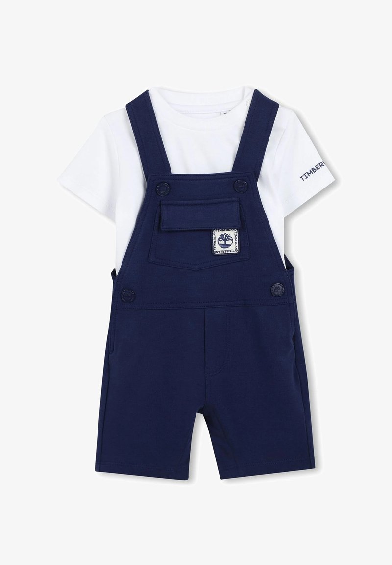 Salopette corta blu navy con tasca frontale e toppa con logo, abbinata a una maglietta bianca. Presenta bretelle regolabili e chiusura con bottoni.