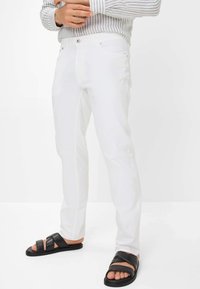 Weiße gerade geschnittene Hosen mit glatter Textur, ausgestattet mit einem Knopfverschluss und kontrastierenden Nähte. Kombiniert mit schwarzen Rutschsandalen.