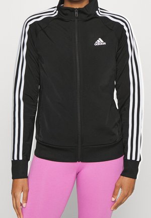Person wearing black Adidas zip-up jacket with white stripes on sleeves and pink leggings, standing with arms relaxed by sides.