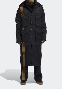 Černá bunda se zlatými pruhy, několika kapsami, patentními uzávěry a vrstveným designem. Má dlouhou siluetu a odpovídající kalhoty.