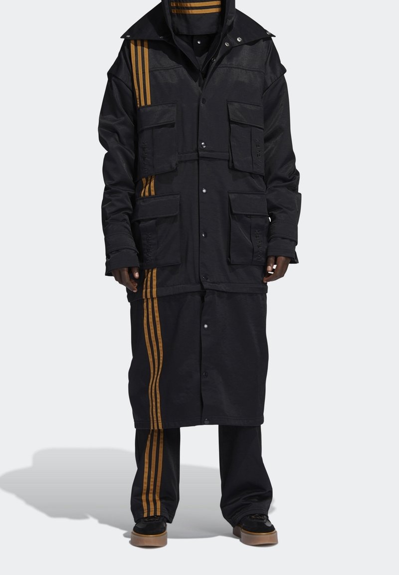 Černá bunda se zlatými pruhy, několika kapsami, patentními uzávěry a vrstveným designem. Má dlouhou siluetu a odpovídající kalhoty.