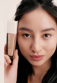 Foundationflaske med et kvadratisk beige lokk, som viser en lys brun væske. Modellen holder flasken nært sit ansigt, med naturlig makeup.