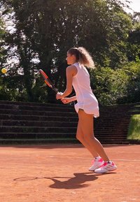 Eine Tennisspielerin in einem weißen Outfit und Schuhen mit pinken Akzenten bereitet sich darauf vor, einen Tennisball auf einem Außenplatz aus rotem Sand zu schlagen, mit Bäumen im Hintergrund.