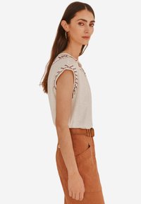 Haut sans manches gris clair avec des détails brodés sur les coutures des épaules, associé à un pantalon taille haute de couleur rouille et une ceinture.