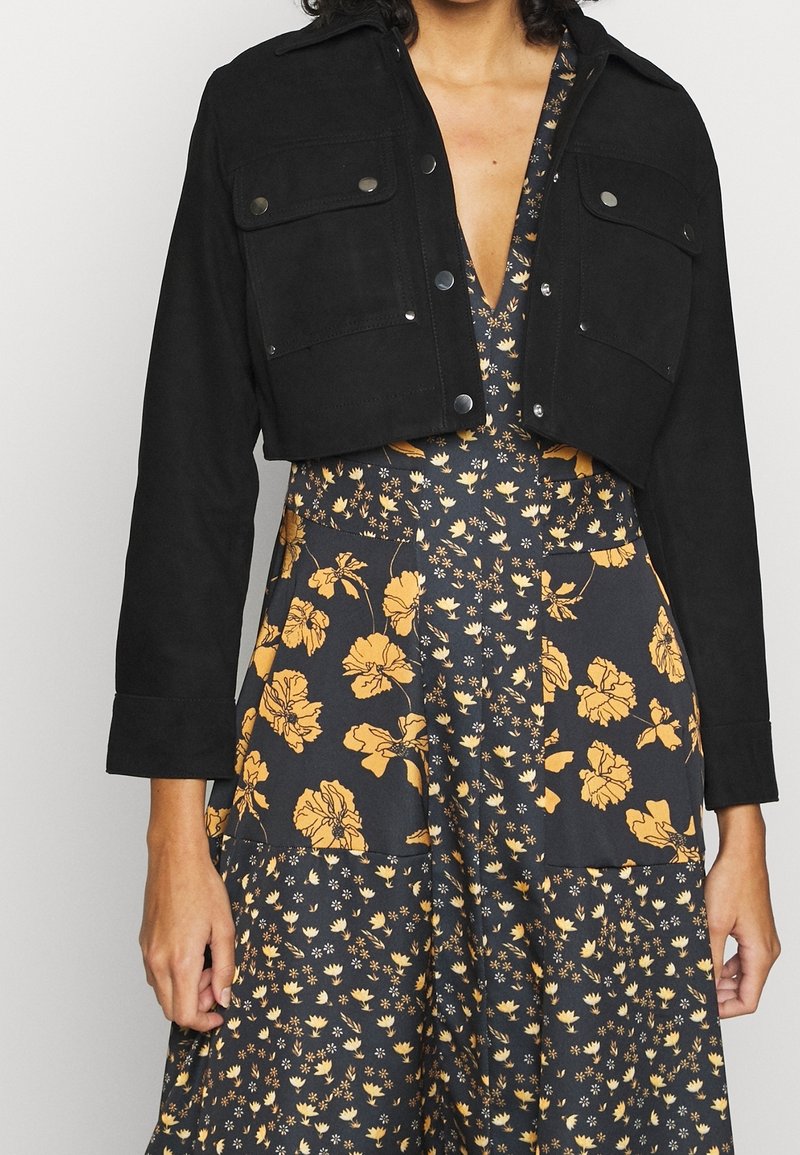 Femme portant une veste noire courte sur une robe à col en V à motif floral noir et jaune, debout devant un fond clair uni.