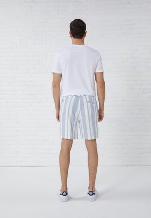 Mężczyzna stoi twarzą do białej ceglanej ściany, ubrany w gładki biały t-shirt, niebiesko-białe szorty w paski oraz białe trampki z granatowymi detalami.