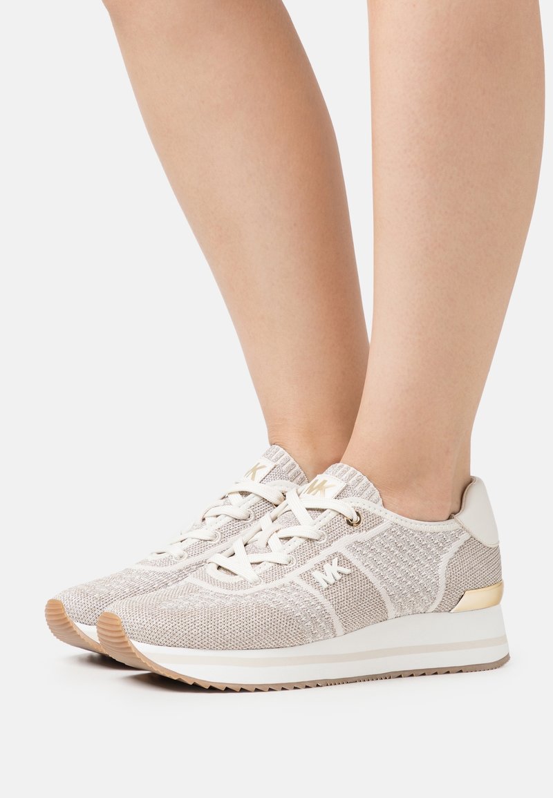 Personne portant des baskets beiges en tricot avec des lacets blancs, des semelles compensées et le logo Michael Kors sur le côté et la languette.