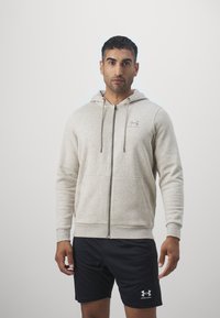 Felpa con zip di colore grigio chiaro, dotata di cappuccio con cordino, tasche frontali e polsini a coste, con un logo sottile sul petto. Pantaloni corti neri.