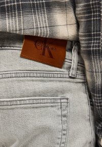 Καφέ δερμάτινη ετικέτα Calvin Klein σε ανοιχτό γκρι τζιν τζιν που φοριέται με γκρι και μαύρο καρο πουκάμισο.