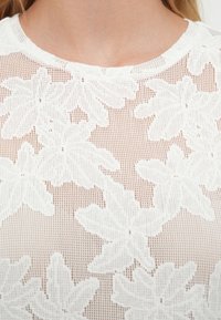 Haut en dentelle florale blanche avec un motif en maille, un col rond, des motifs délicats en forme de feuilles et une finition texturée.