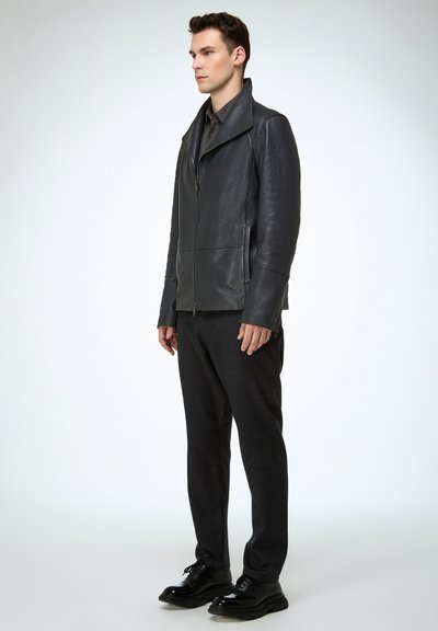 Schwarze Lederjacke mit hohem Kragen, Reißverschluss und zwei Vorderfachern. Kombiniert mit dunkler Hose und glänzenden schwarzen Schuhen.