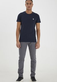 Jūras zils īsām piedurknēm t-krekls ar mazu kvadrātveida logotipu, apvienots ar gaiši pelēkiem slaidiem biksēm un melniem sporta apaviem, attēlots priekšā.