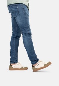 Beige Sneakers mit grünen und braunen Akzenten, strukturiertem Gummisohle, kombiniert mit blauen engen Jeans. Fokus auf Schuhdesign und Materialien.